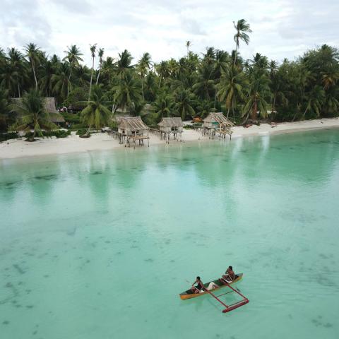 Little native wood boat with two indigenous fishermen paddling in the green sea with beach an palms in background