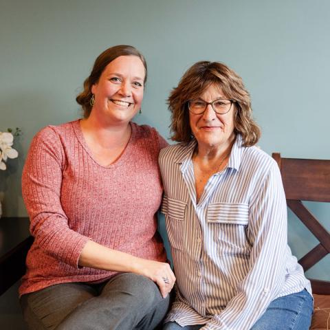 Hillary Hauser and Karina Johnston sitting closely side by side on wooden bench in room with gray background