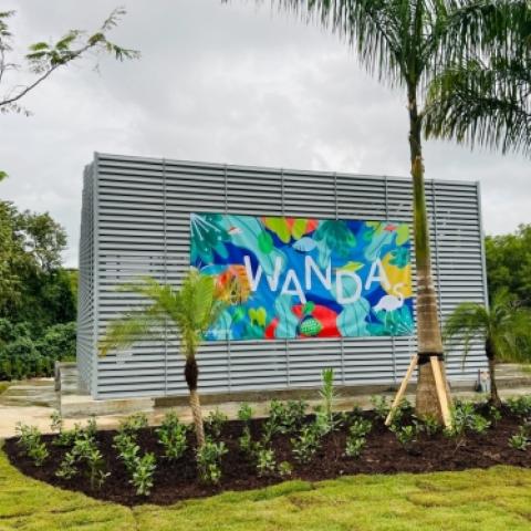 A steel structure sits behind palm trees and is adorned with a bright, multicolored banner reading "Wanda"