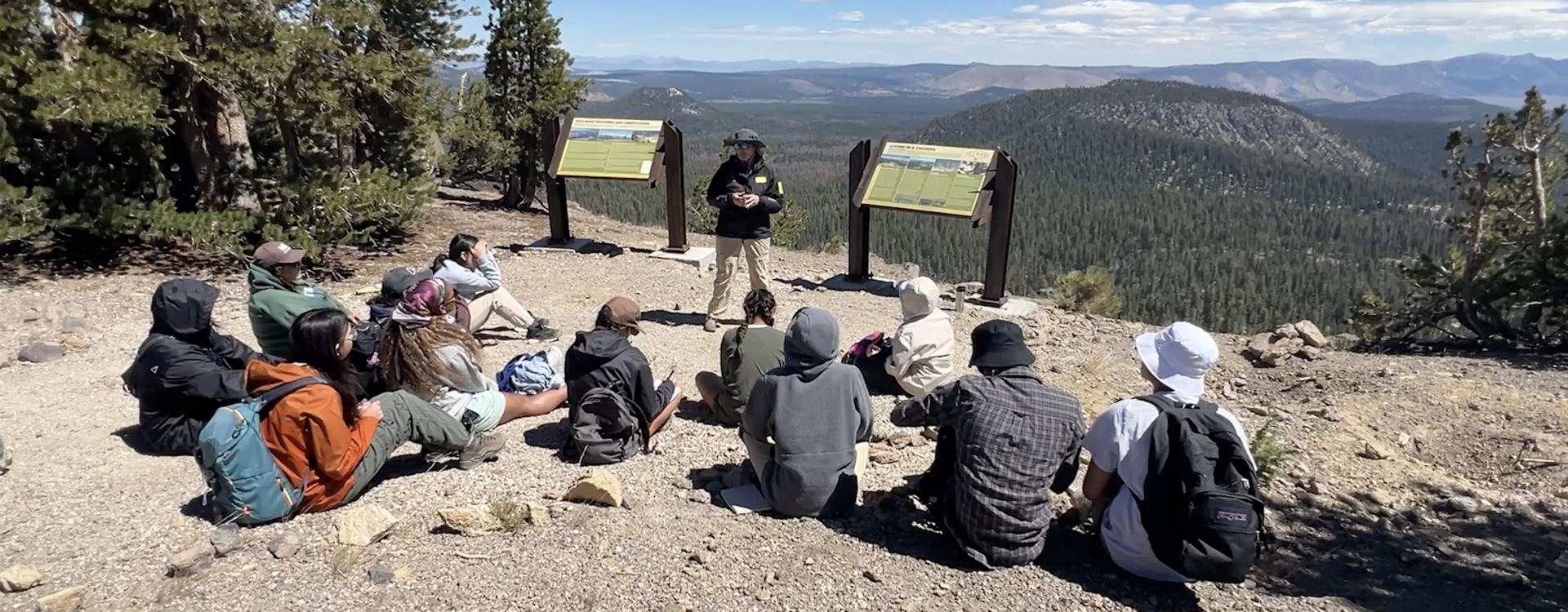 FUERTE group of students sitting on ground listening to docent standing against stunning mountain background view