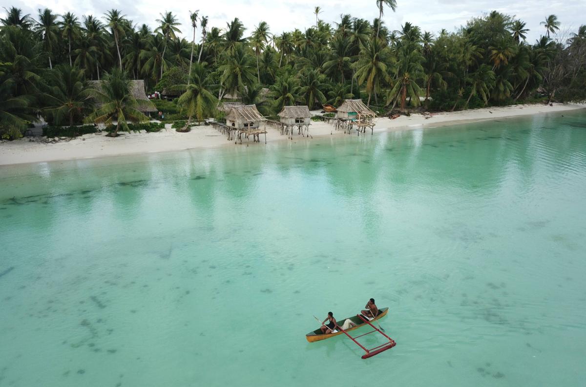 Little native wood boat with two indigenous fishermen paddling in the green sea with beach an palms in background