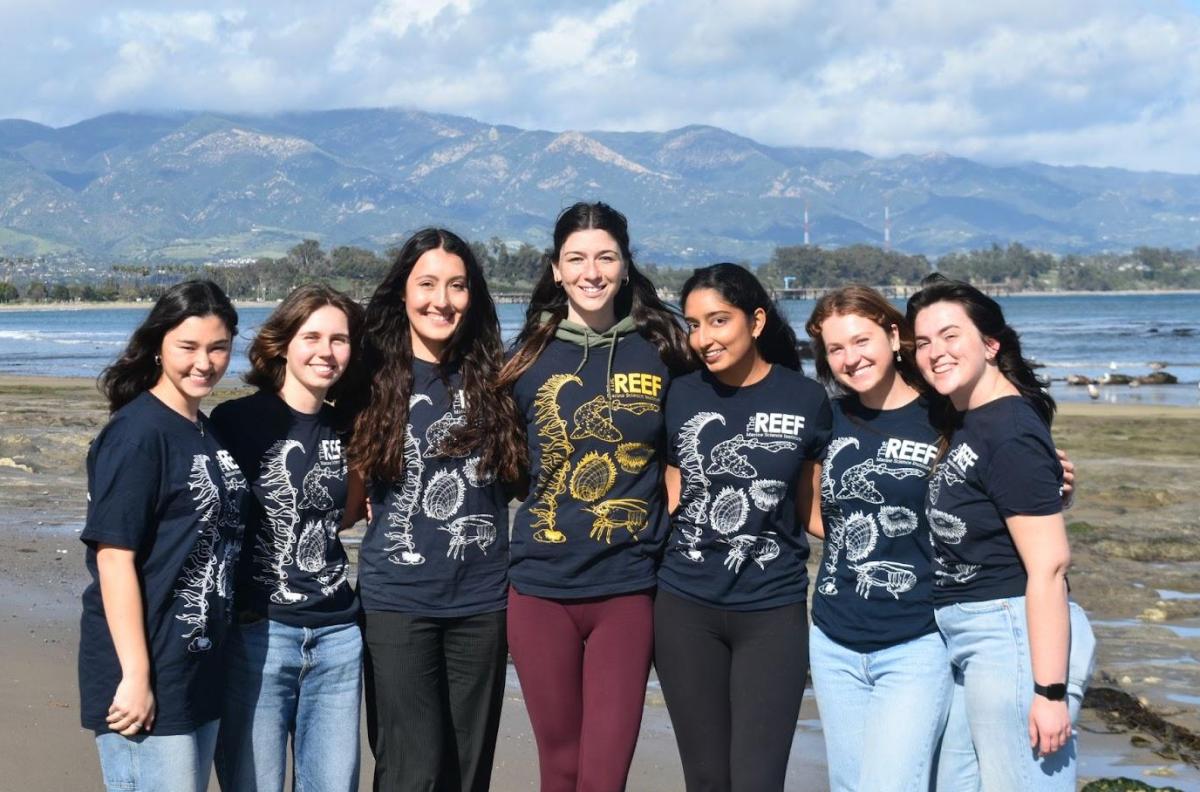 Group of seven students wearing REEF t-shirt posing on Campus Point beach with mountain background