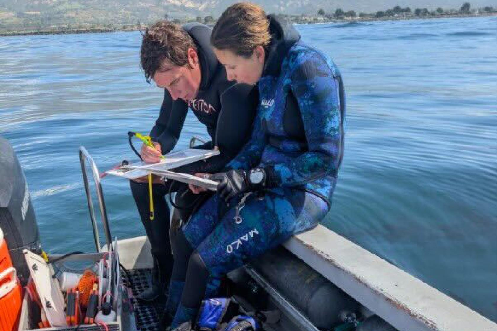 Avery on left of research partner consulting notes sitting on edge of small boat and wearing wetsuits