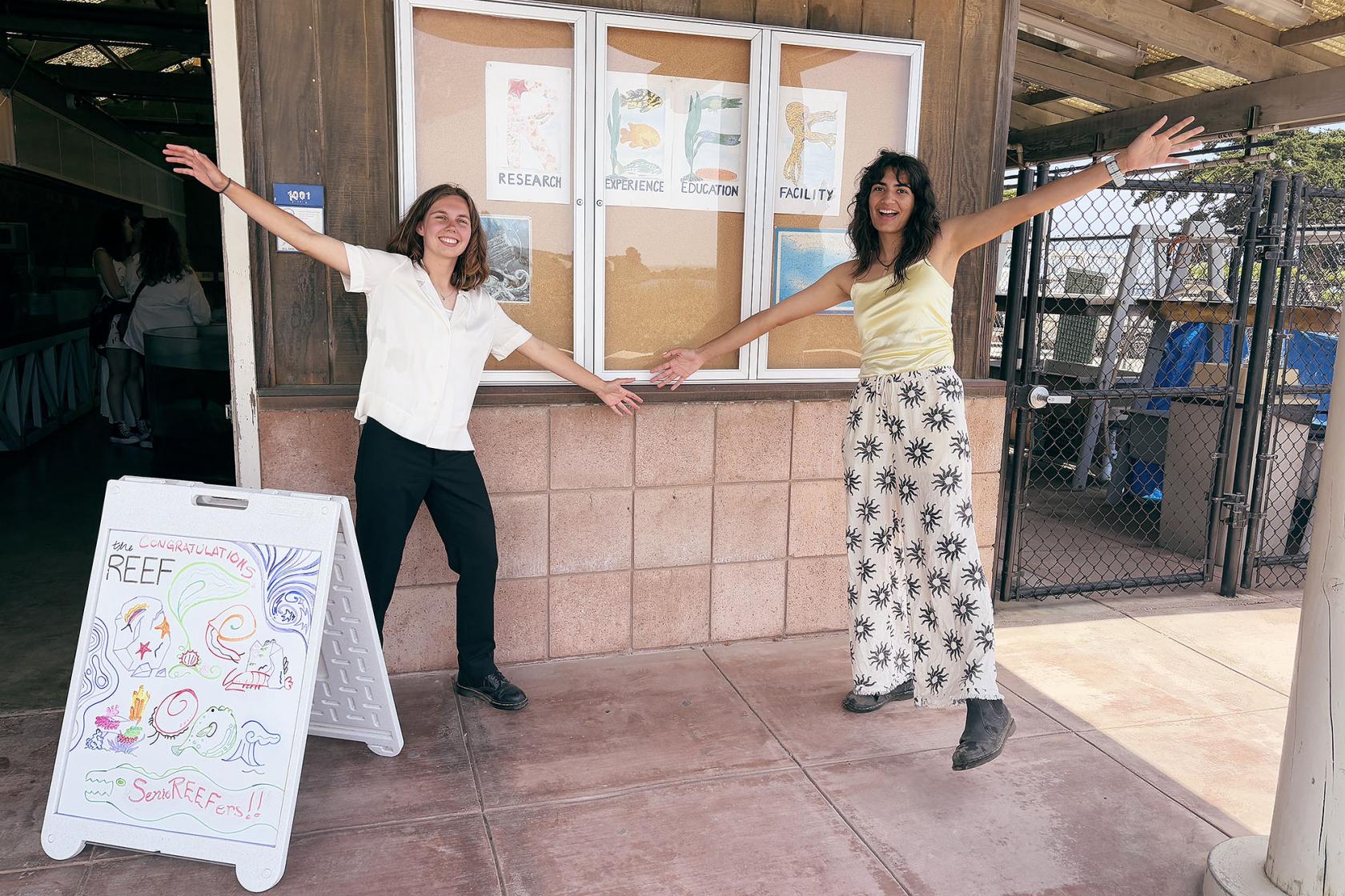 Marine and Gianna standing in front of the REEF, pointing at poster on wall